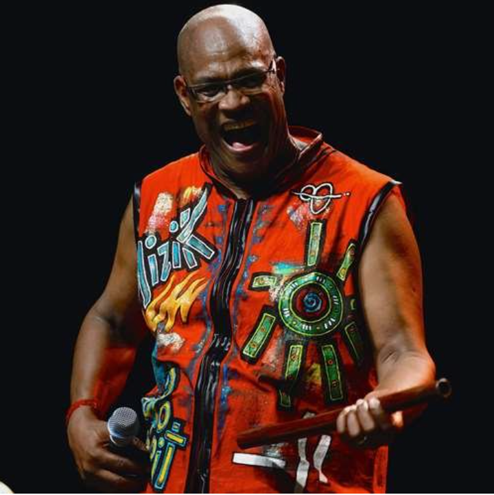 Le musicien Dédé en pleine performance sur scène. Il porte un gilet rouge vif peint à la main par l'artiste Baboo, tient un micro et une flûte en bois, souriant. Artiste et musicien Zouk/Caraïbes.